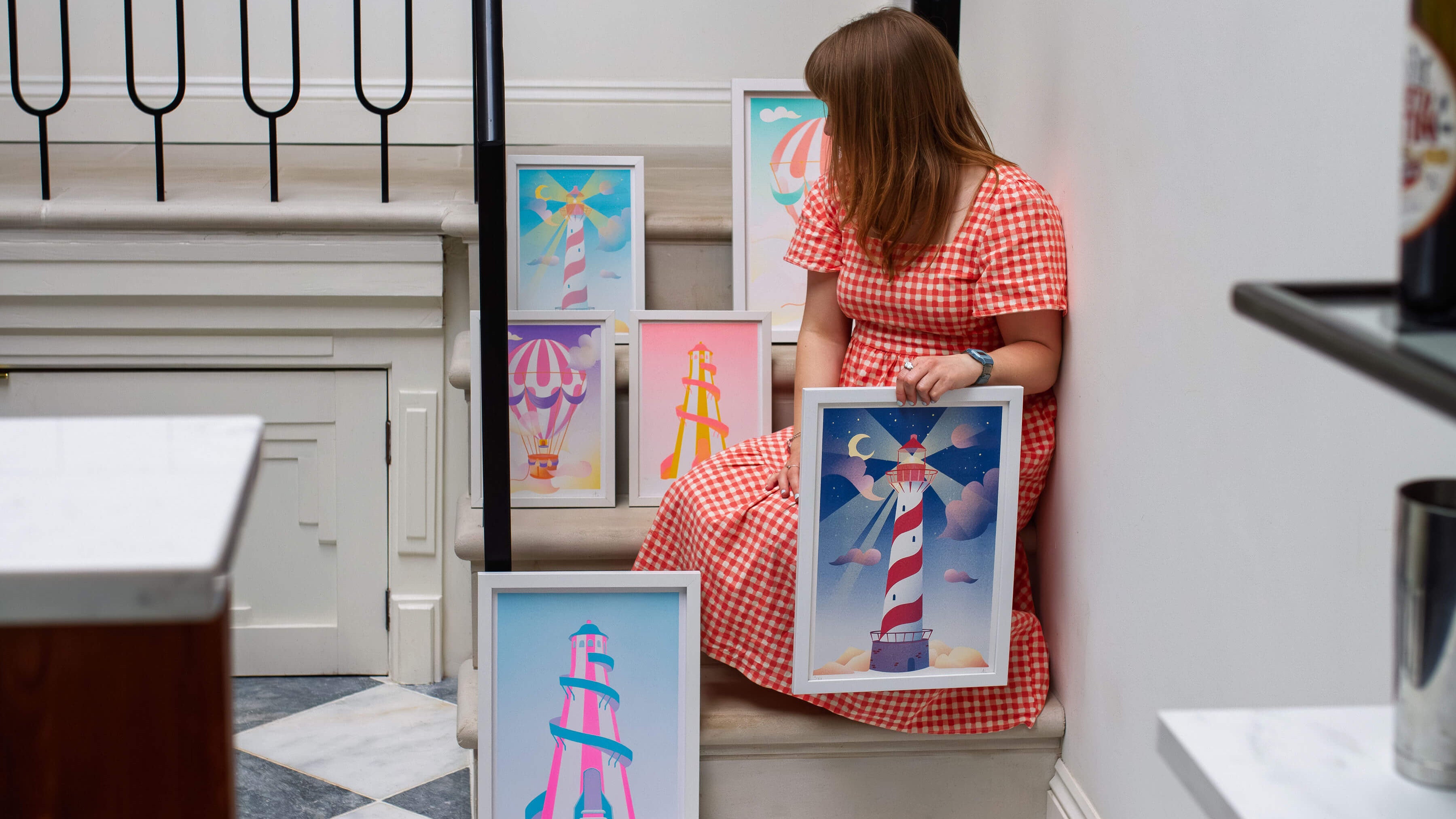 Woman holding a framed artwork of a Lighthouse, sitting on steps surrounded by more risographs prints of hot air balloons and helter-skelters.